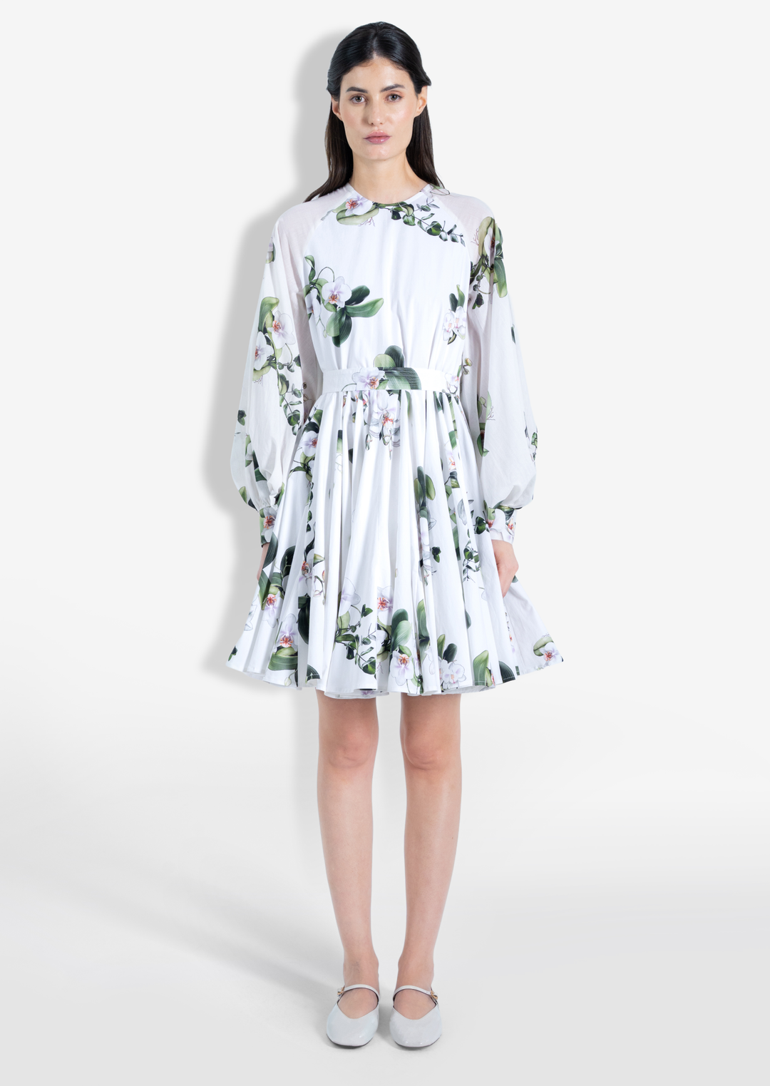 A model wearing a white short dress with an orchid floral print, long ruffled sleeves, and a flared silhouette. The dress has a concealed back zipper.