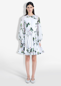 A model wearing a white short dress with an orchid floral print, long ruffled sleeves, and a flared silhouette. The dress has a concealed back zipper.