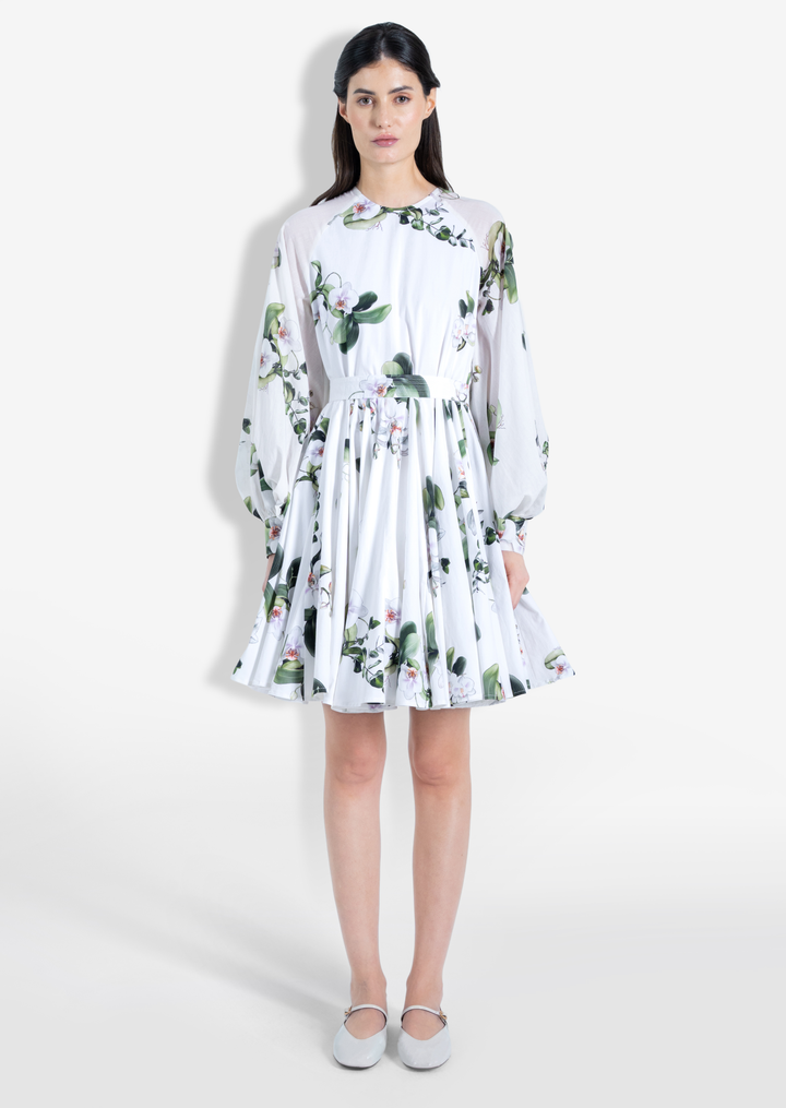 A model wearing a white short dress with an orchid floral print, long ruffled sleeves, and a flared silhouette. The dress has a concealed back zipper.